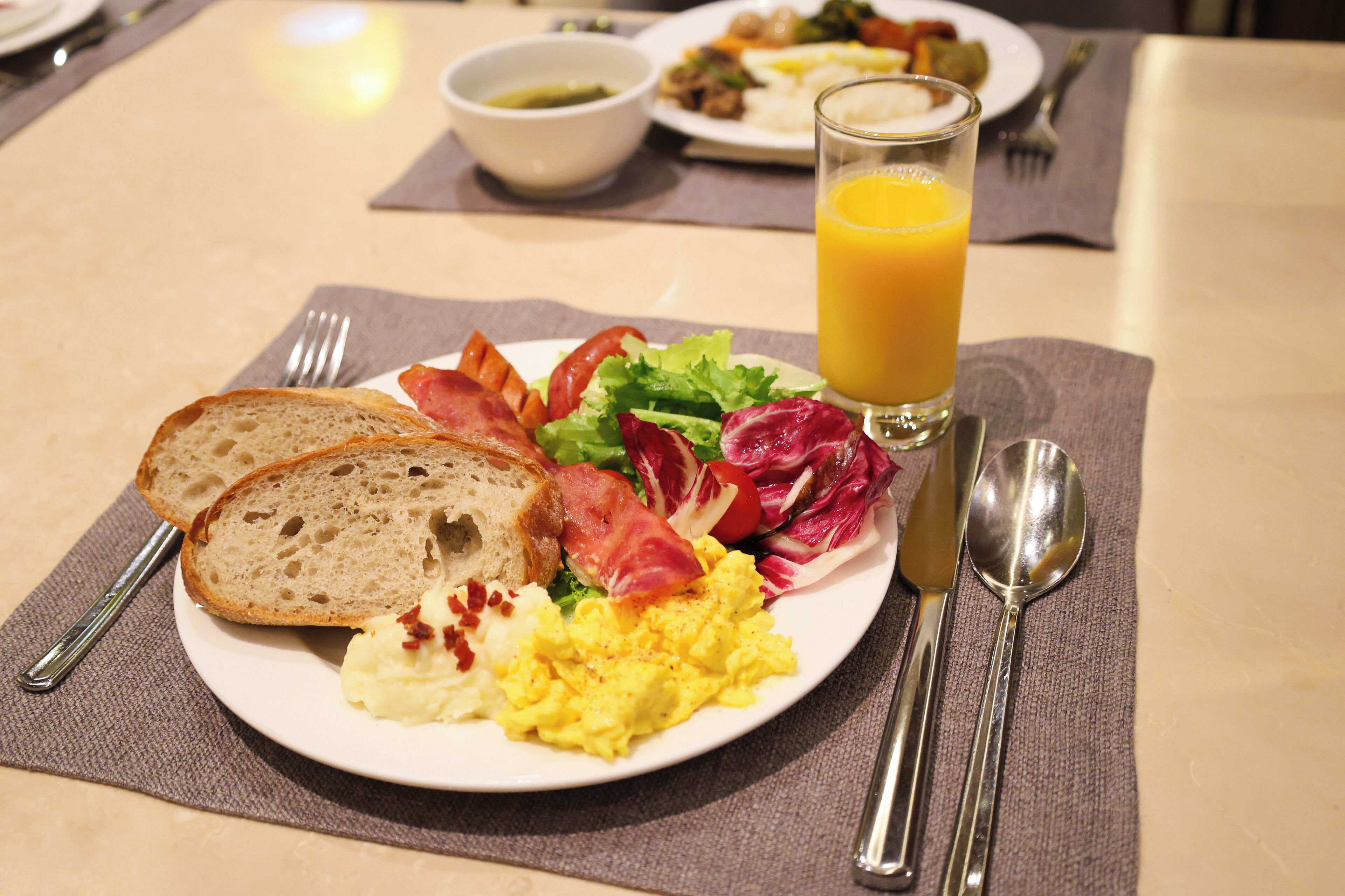 A breakfast plate with toast, scrambled eggs, bacon, and salad, a glass of orange juice, and cutlery on a table.