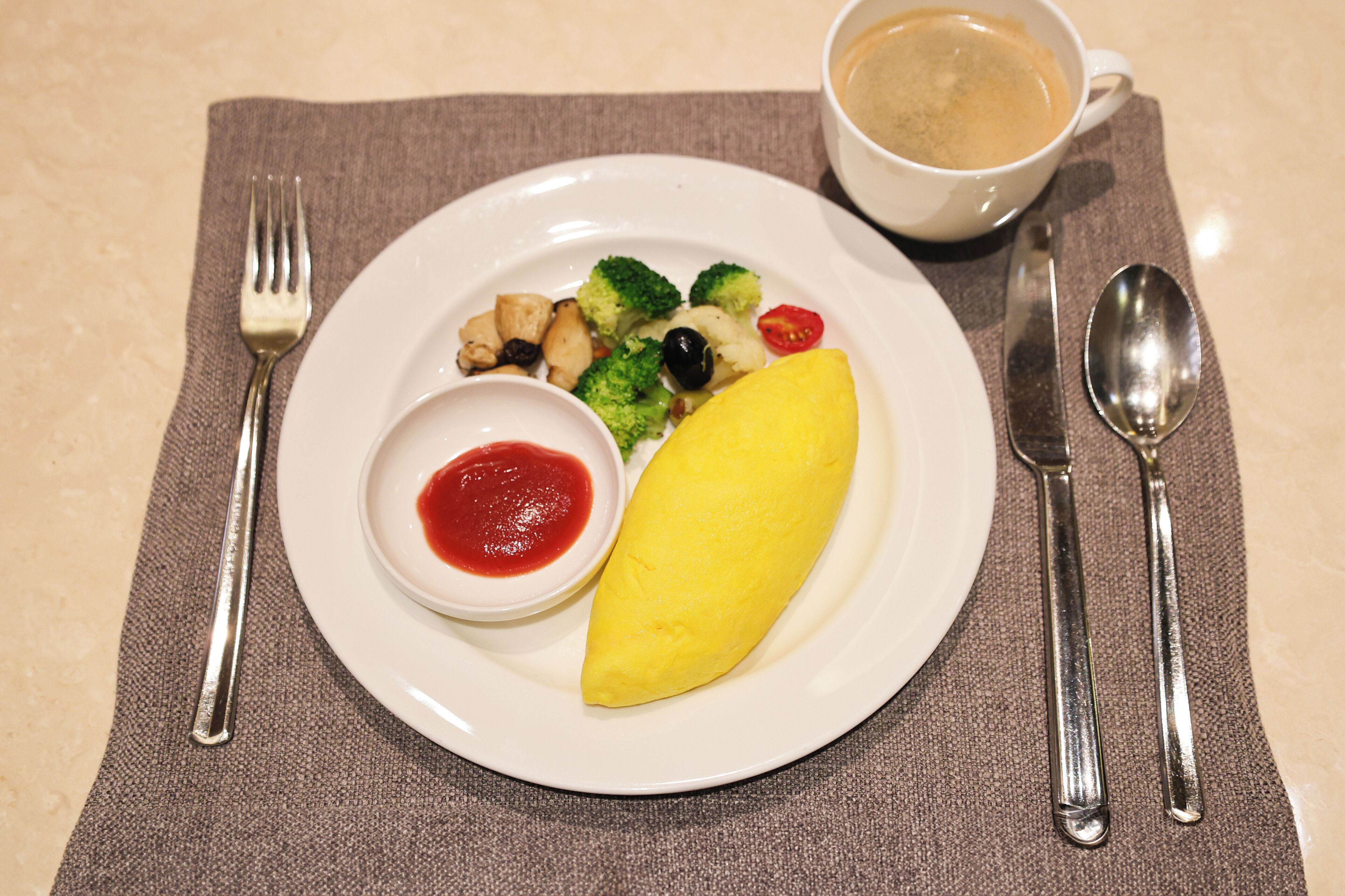 Une assiette contenant une omelette jaune, des brocolis, des tomates cerises et un petit bol de ketchup, accompagnée d’une tasse de café posée sur une nappe.