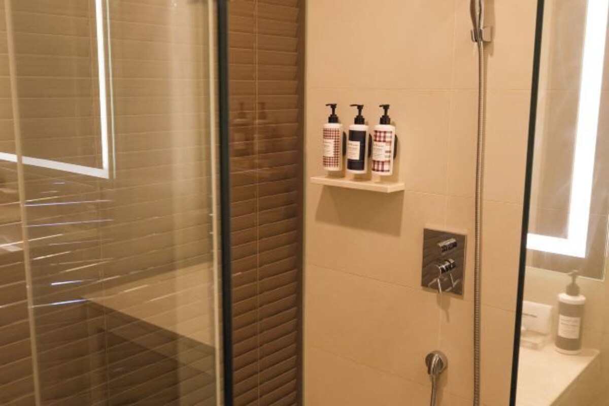 Une douche fermée par des parois de verre, avec un mur recouvert de carreaux, une étag ère pour les articles de toilette et un panneau de commande.