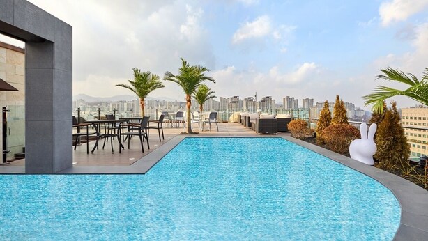 A rooftop swimming pool with lounge chairs and palm trees, overlooking a cityscape. - Skypark Central Seoul Pangyo (Seongnam)