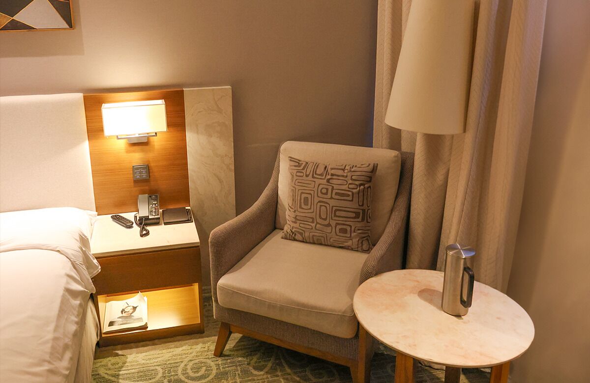 Habitación de hotel con una cama, una mesita de noche con lámpara, una silla con cojín estampado y una pequeña mesa con jarra.