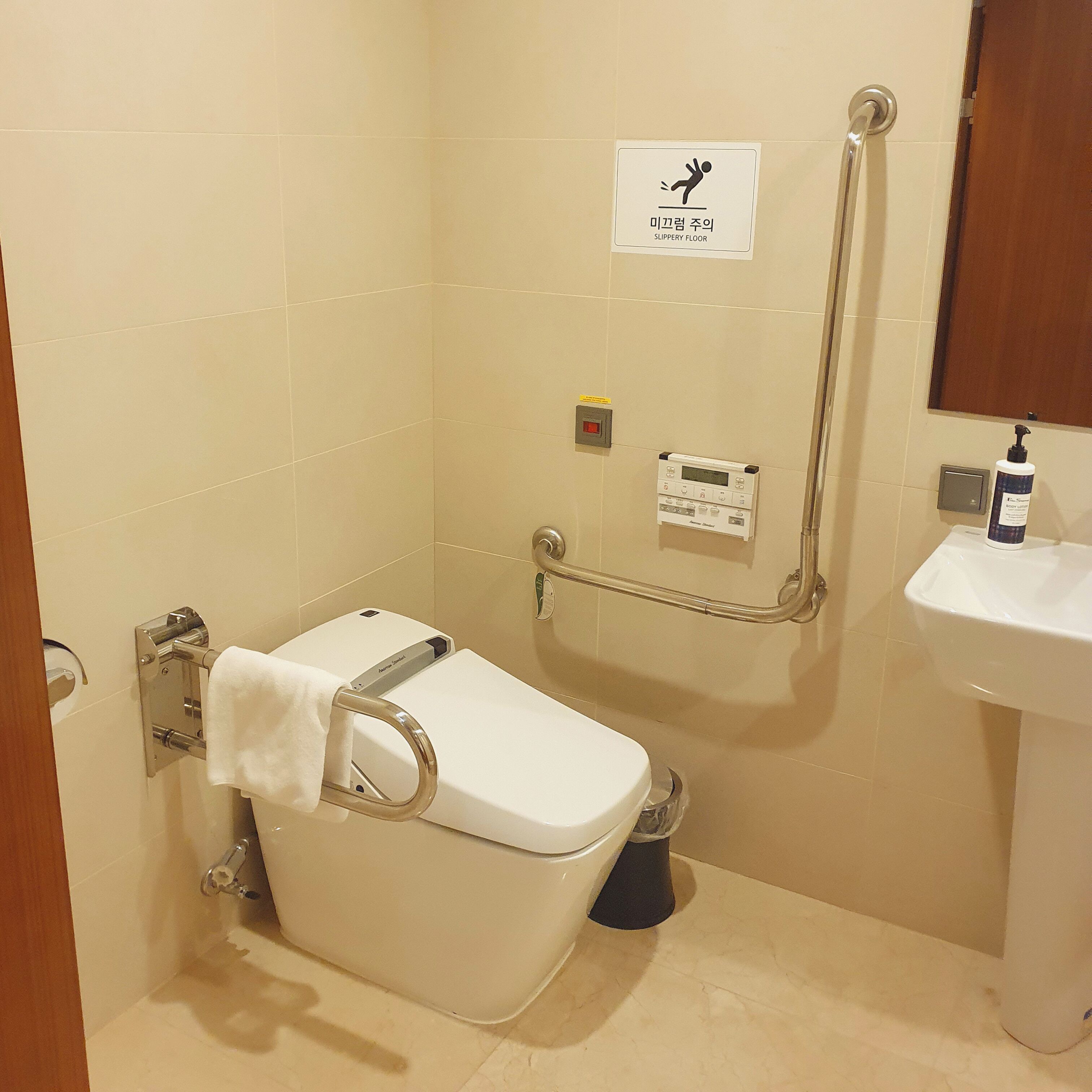 Un baño con inodoro, barra de apoyo, lavamanos y señalización de suelo resbaladizo.