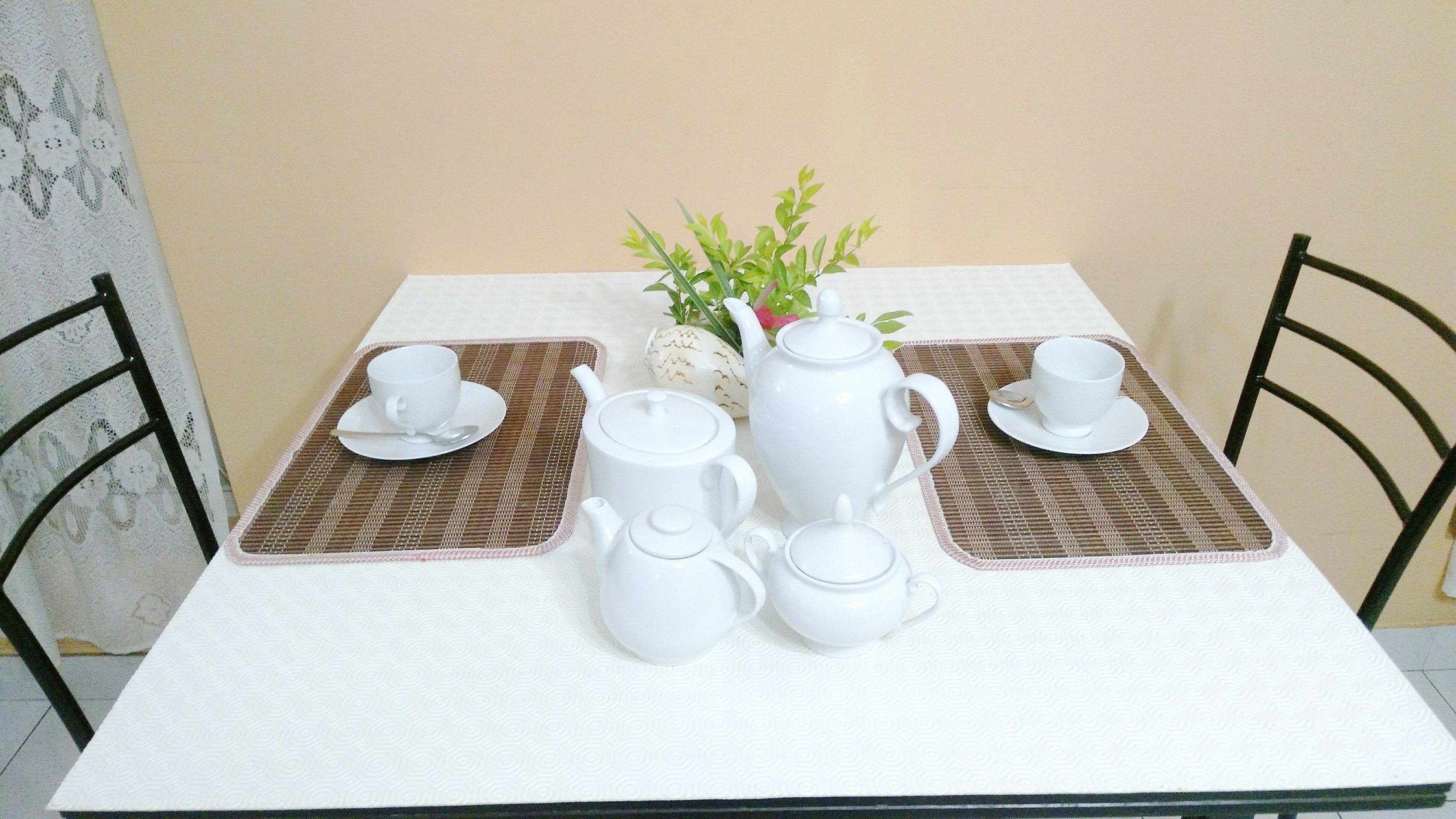 Une table dressée avec des tasses et des soucoupes à thé blanches, une théière et un sucrier, le tout posé sur une nappe blanche agrémentée de sets de table marron.