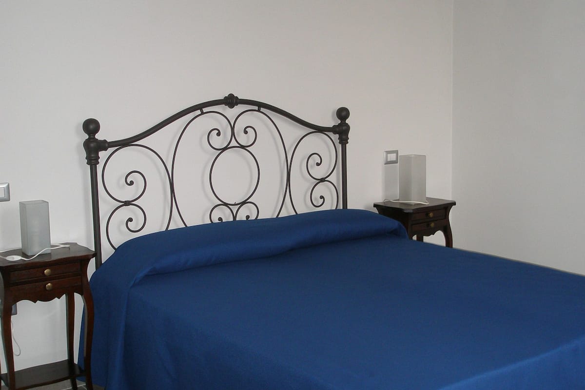 Un dormitorio con estructura de cama metálica, colcha azul, dos mesitas de noche con lámparas y un candelabro.