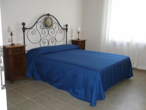Cama bem arrumada com colcha azul, cabeceira de metal com espelho, duas mesas de cabeceira com abajures e piso de azulejos.