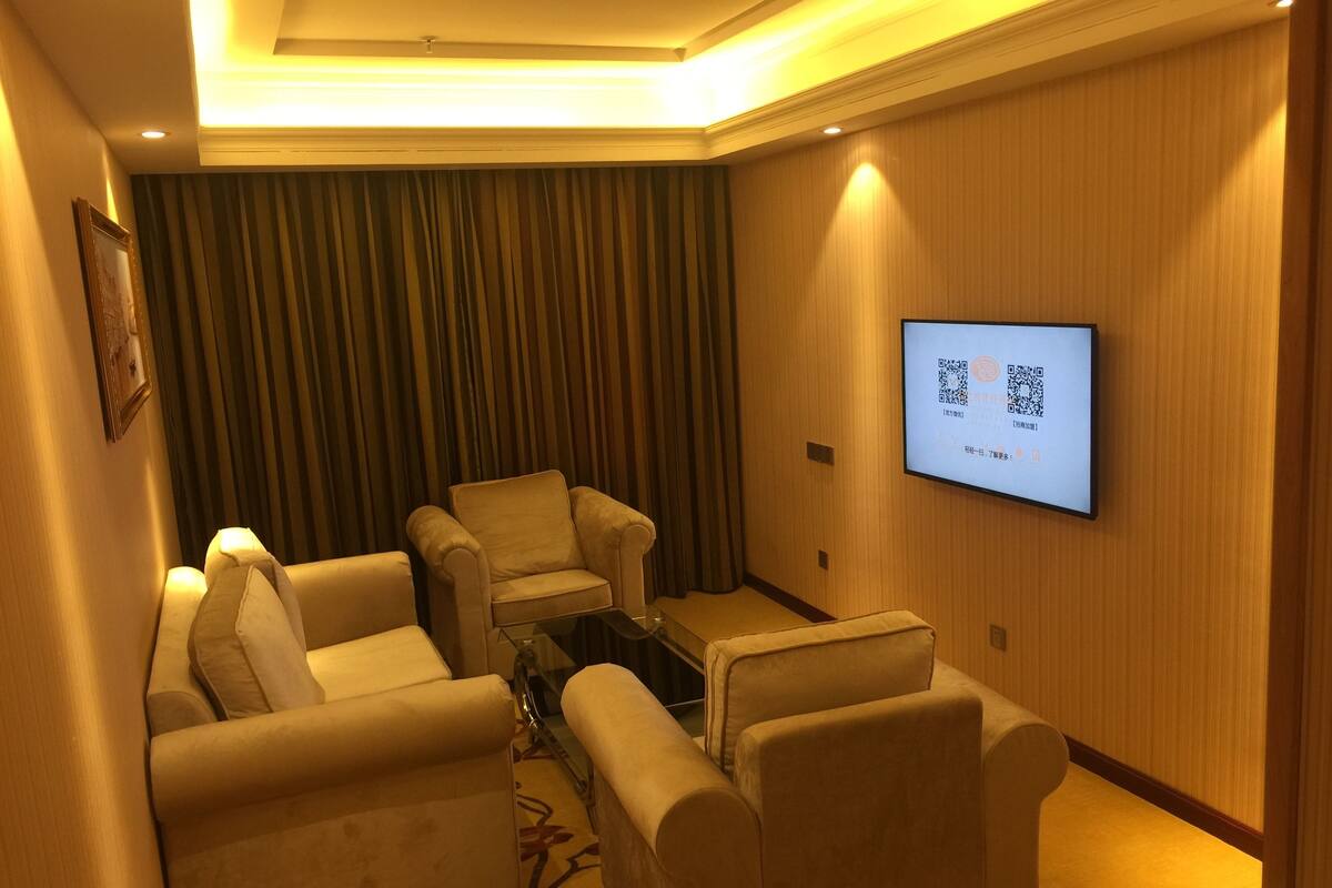 A hotel room with a seating area featuring three sofas and a coffee table, a flat-screen TV mounted on the wall, and a curtain backdrop.