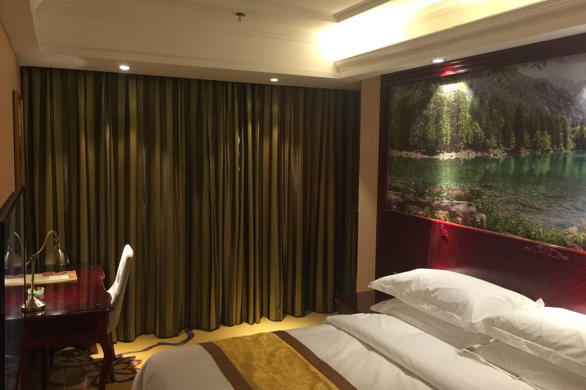 A hotel room with a large bed, a desk with a lamp, a trash bin, and a painting of a forest scene.