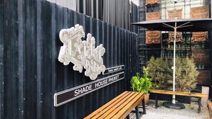Pared corrugada negra con un letrero que dice 'SHADE HOUSE PHUKET' y un banco con plantas en macetas.
