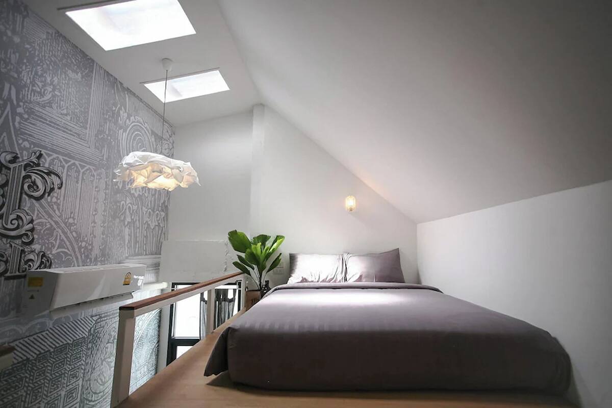 Quarto com cama, escrivaninha, cadeira, planta e janela.