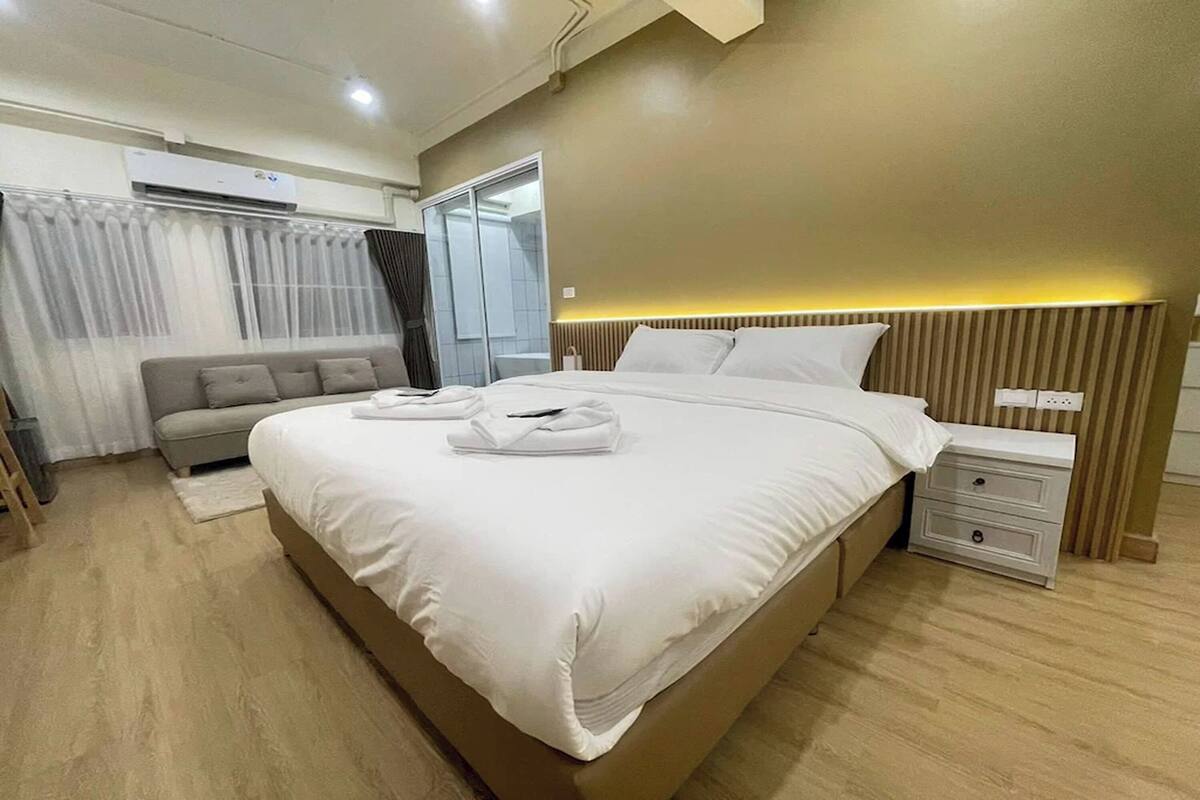 Uma cama bem arrumada com lençóis brancos, uma mesa de cabeceira com abajur, um sofá e um guarda-roupa com portas deslizantes.
