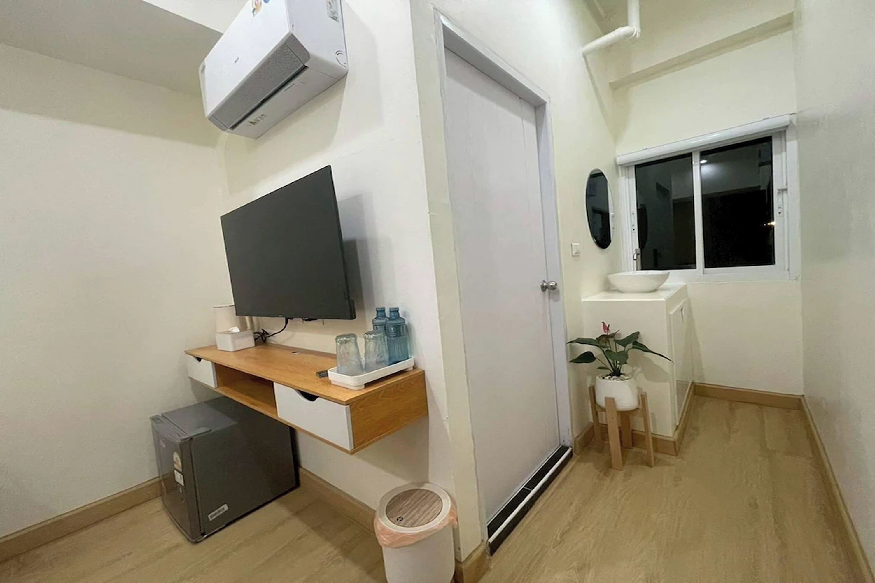 A room with a flat-screen TV, a small wooden desk, a radiator, a window, and a potted plant.
