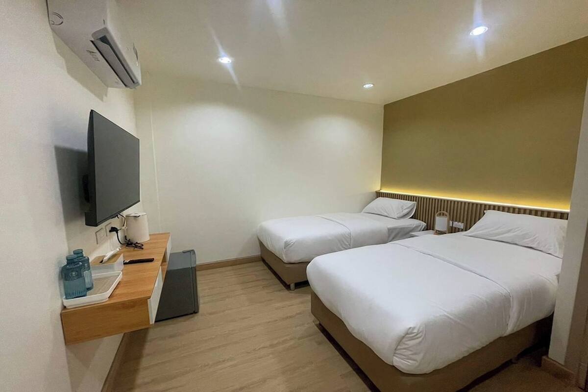 Quarto de hotel com duas camas, uma televisão, uma escrivaninha com abajur e aparelho de ar condicionado.