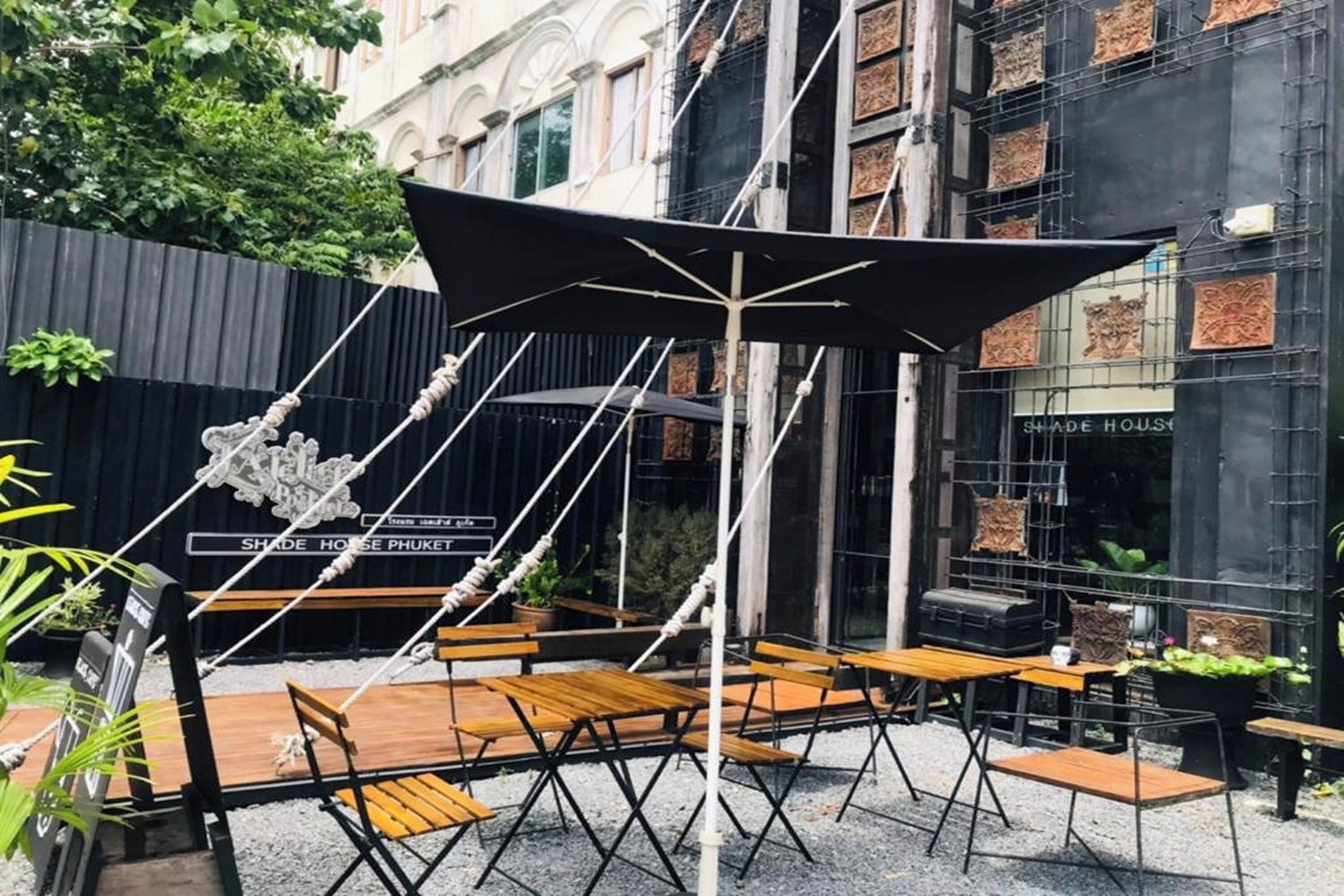 An outdoor seating area with wooden tables and chairs, a large black umbrella, and a building with 'SHADE HOUSE' signage in the background.