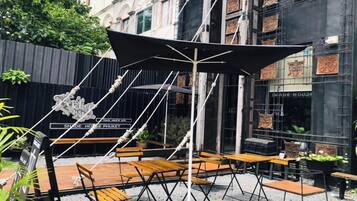 An outdoor seating area with wooden tables and chairs, a large black umbrella, and a building with 'SHADE HOUSE' signage in the background.