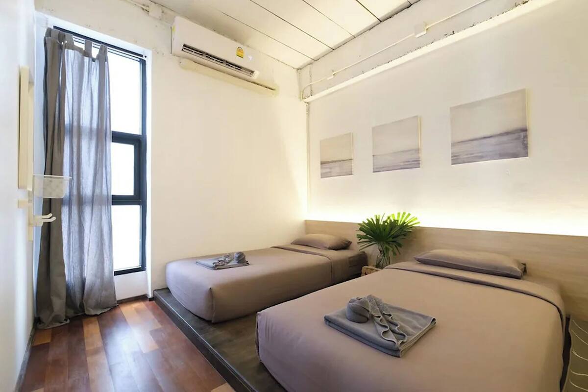 Quarto de hotel com duas camas, janela com cortinas, uma planta e obras de arte na parede.