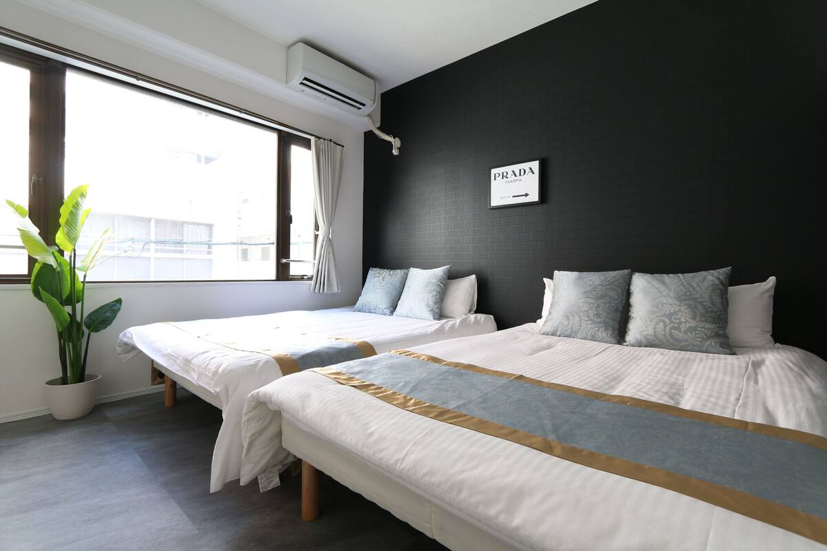 A hotel room with two beds, a potted plant, a window with curtains, and a wall-mounted air conditioner.