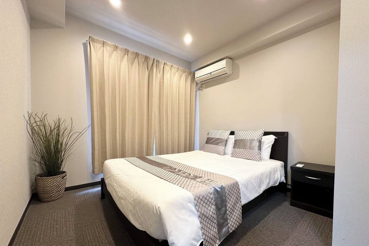A hotel room with a bed, a nightstand, a potted plant, a window with curtains, and an air conditioning unit.