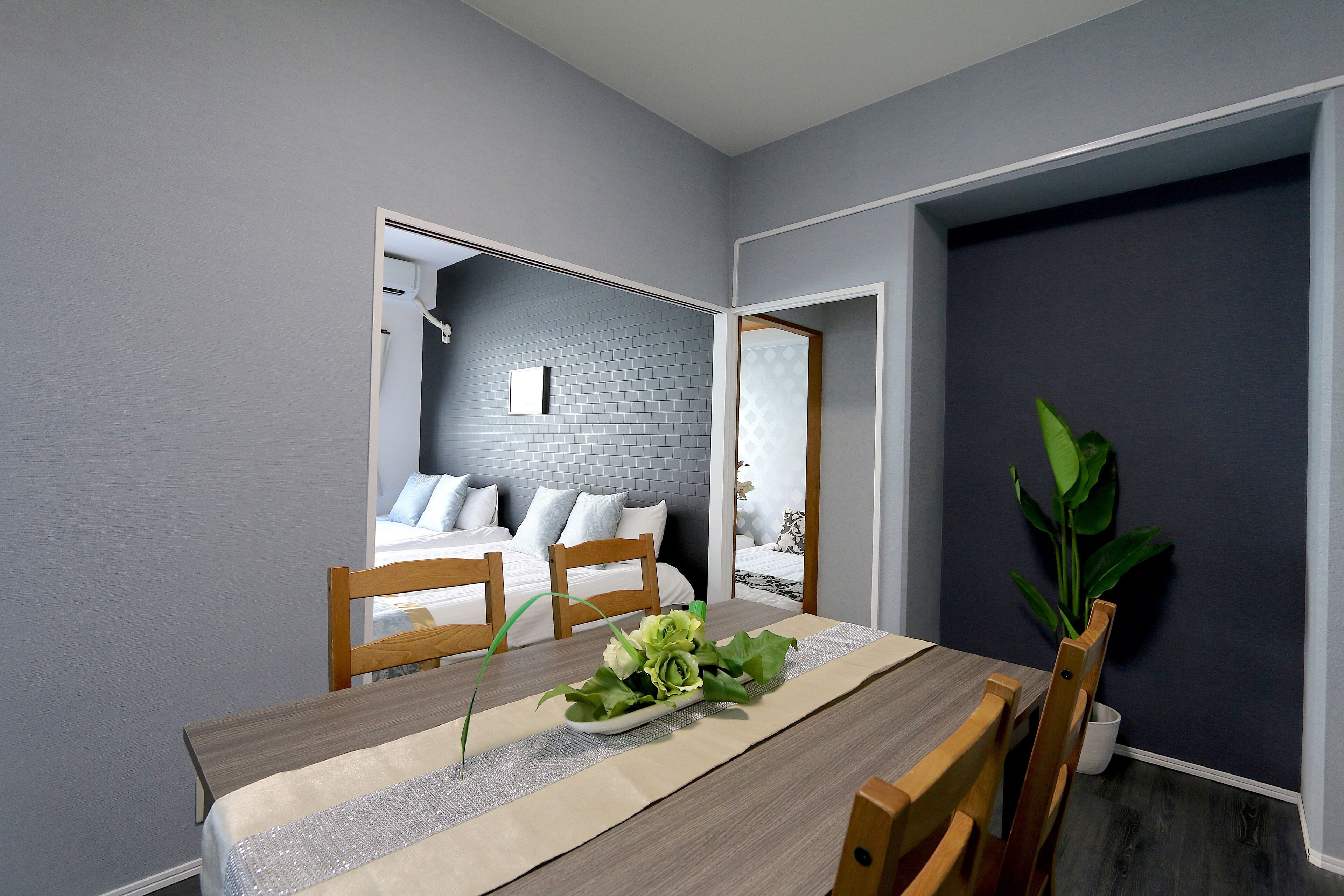 A dining area with a wooden table, chairs, and a vase of flowers. A bed with white bedding is visible in the background.