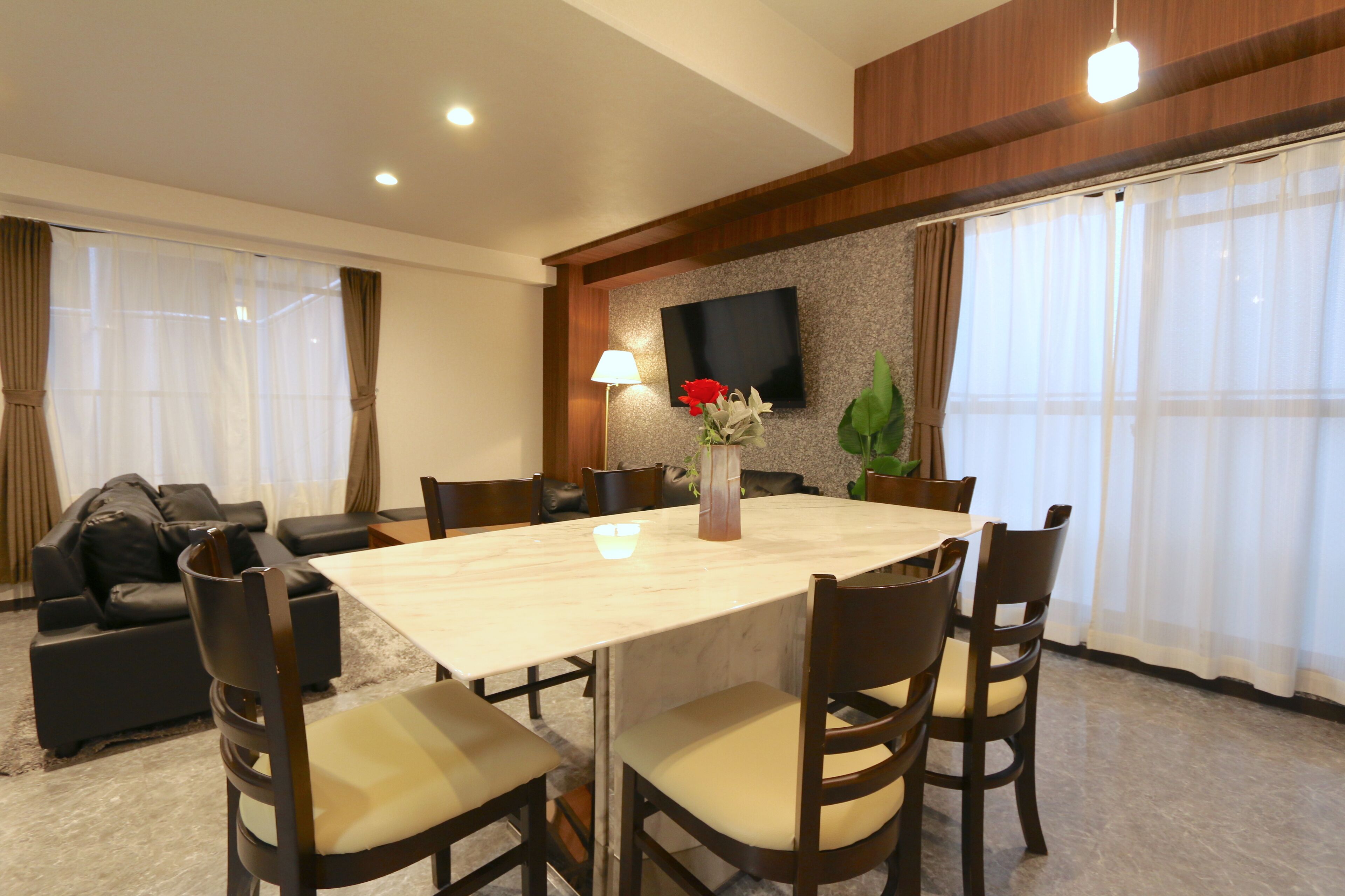 A hotel dining area with a large table, chairs, a sofa, and a TV on the wall.