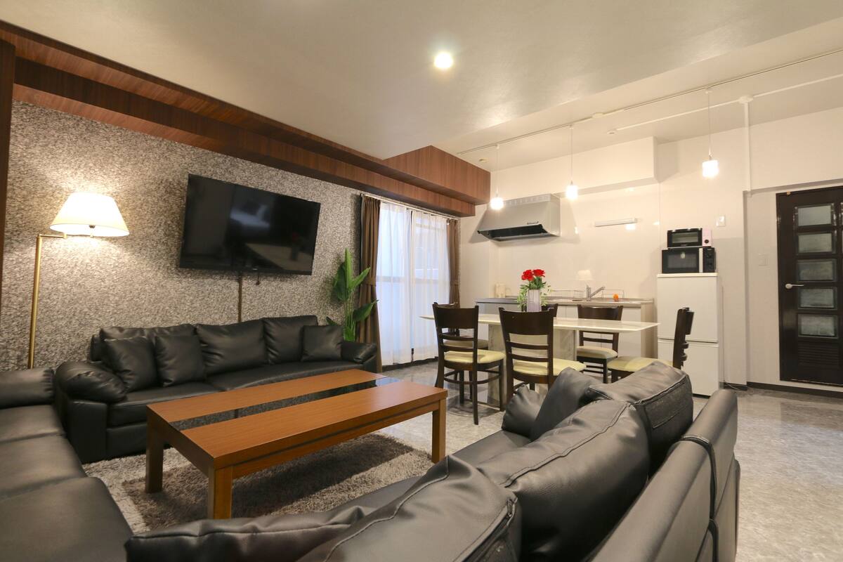 A modern living room with a sofa, a wooden coffee table, and a flat-screen TV mounted on the wall. There is a dining area with a table and chairs, and a kitchen area with a microwave and oven.