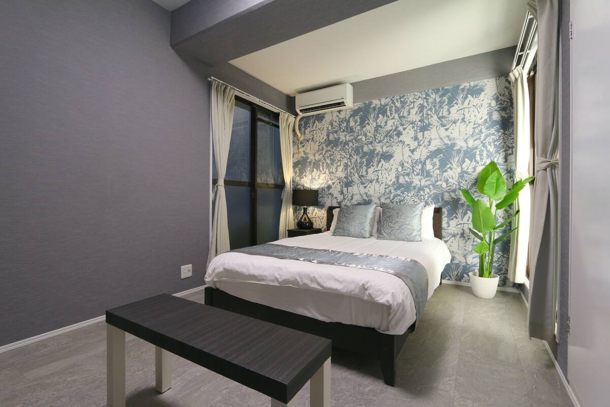 A bedroom with a bed, bedside table, air conditioning unit, curtains, a potted plant, and a patterned wall.