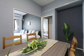 A dining area with a wooden table, chairs, and a vase of green flowers. A bed with white bedding is visible in the background. - Elizabeth Garden (Osaka)