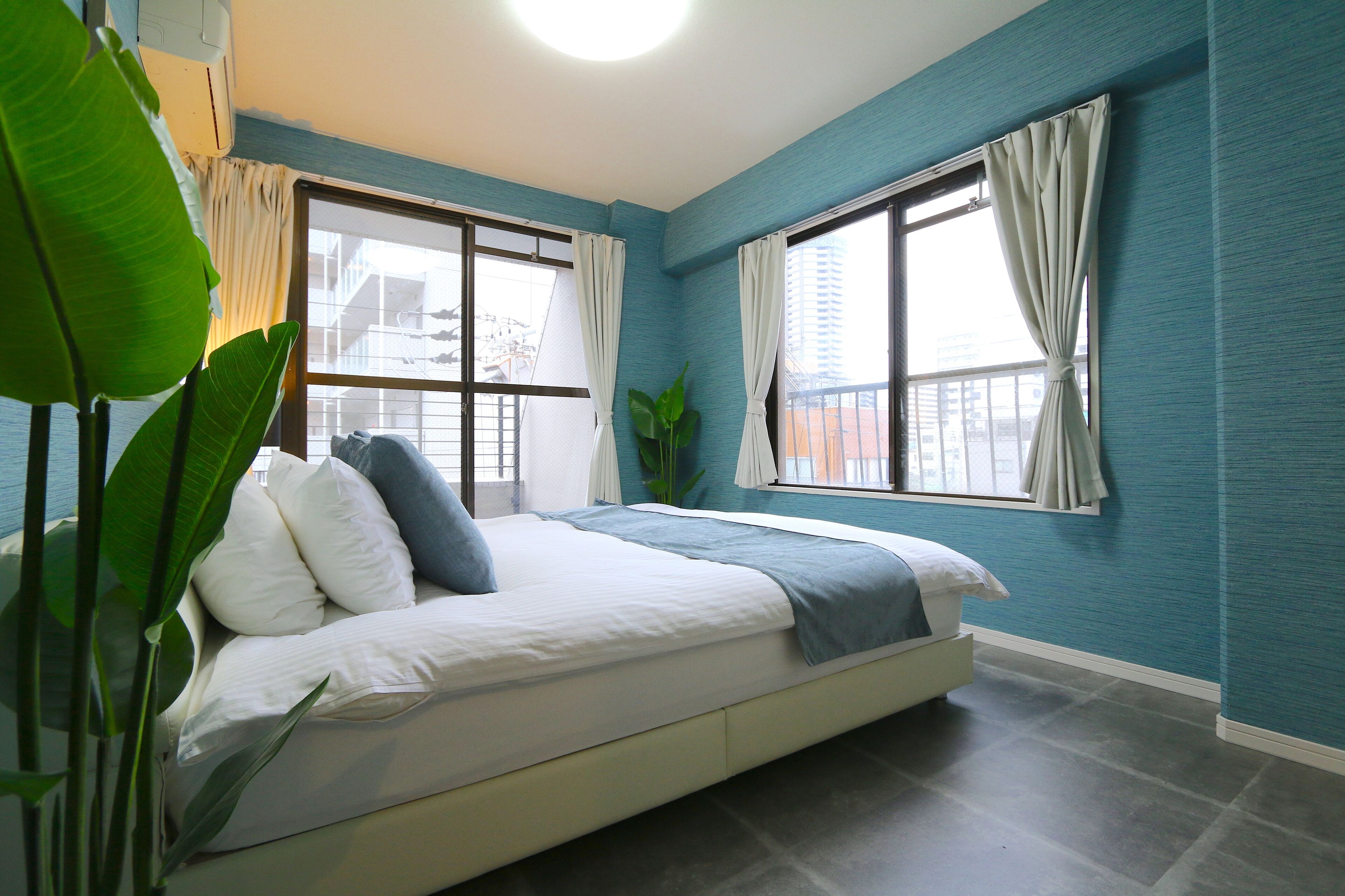 Un dormitorio con una cama, dos ventanas con cortinas, una planta en maceta y vistas de edificios.