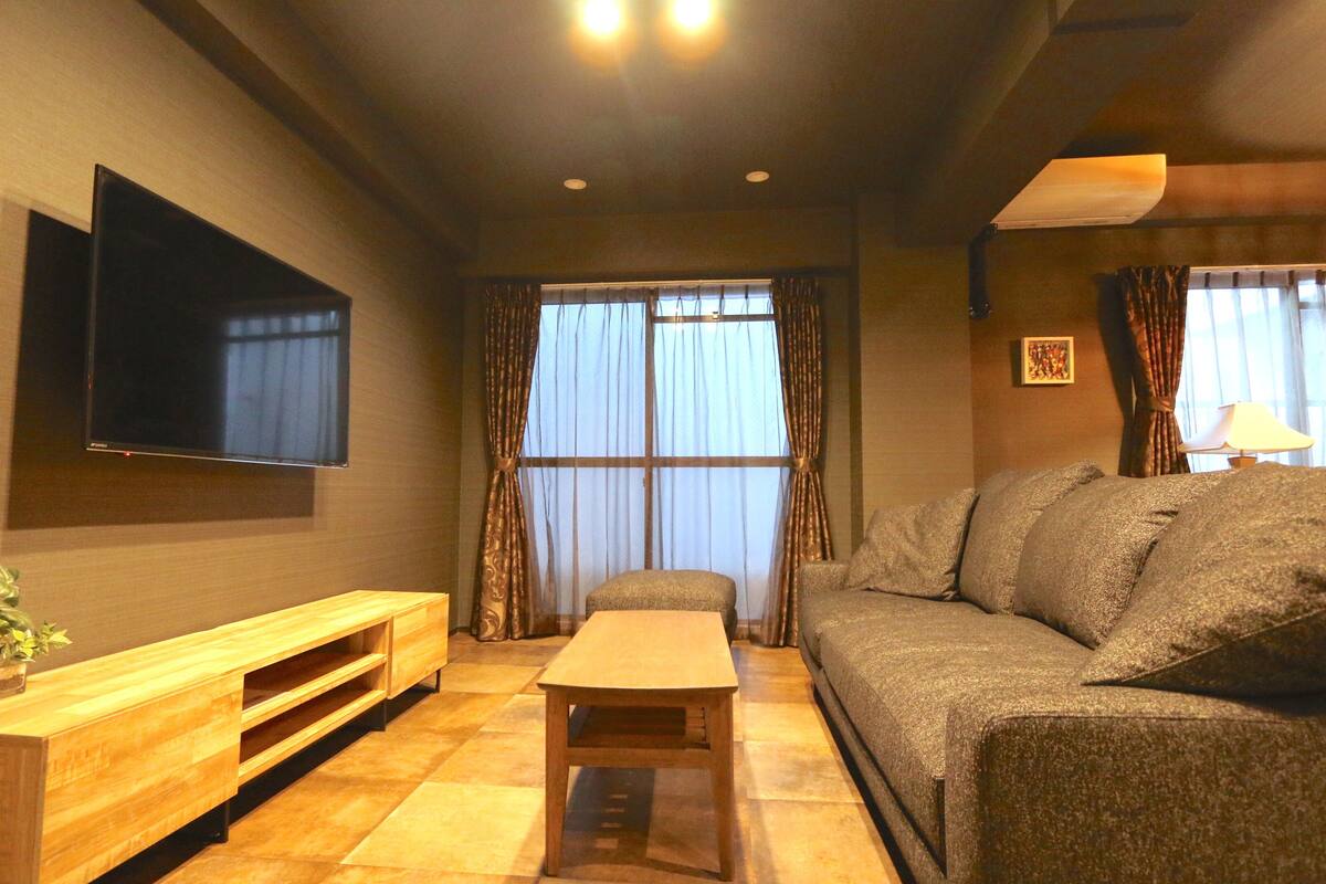 A living room with a sofa, a wooden coffee table, a TV mounted on the wall, and a window with curtains.