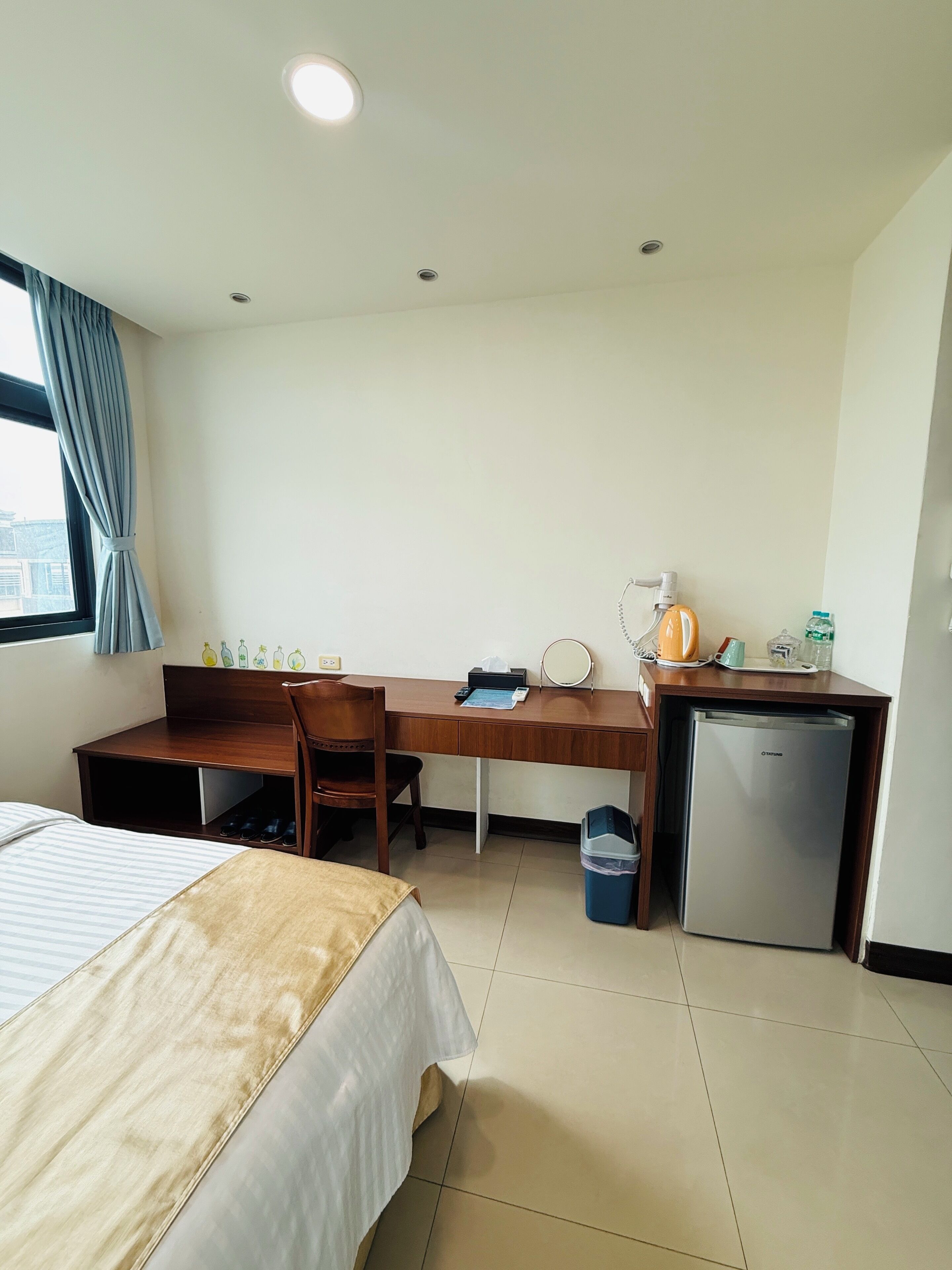 A hotel room with a bed, a desk, a chair, a small refrigerator, and a window with curtains.