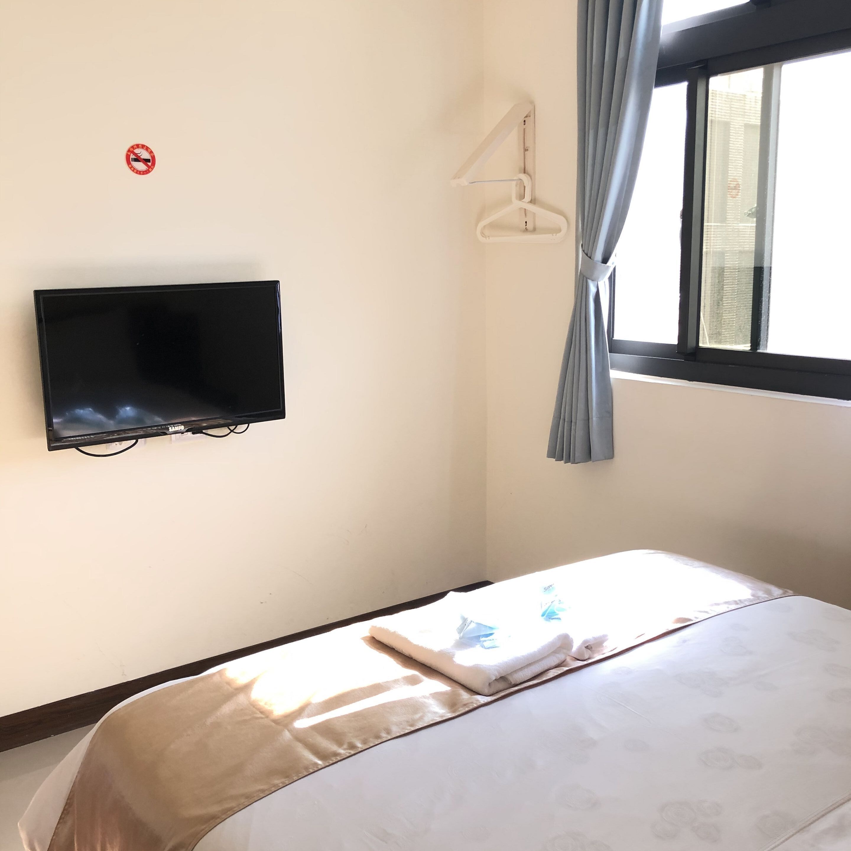A bedroom with a bed, a TV mounted on the wall, a window with curtains, and a no-smoking sign.