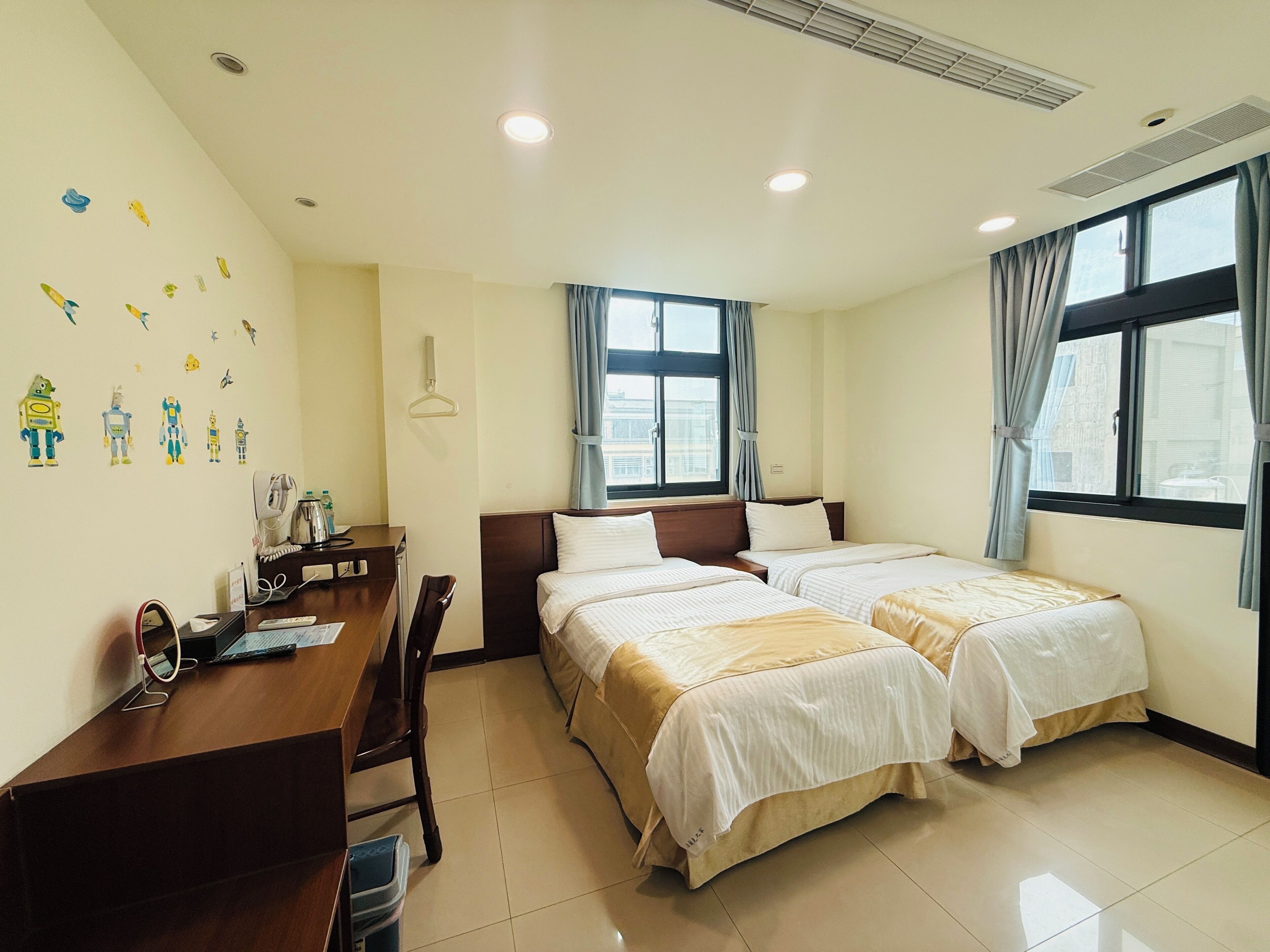 A hotel room with two beds, a desk, and a chair. There are wall decorations and a window with curtains.