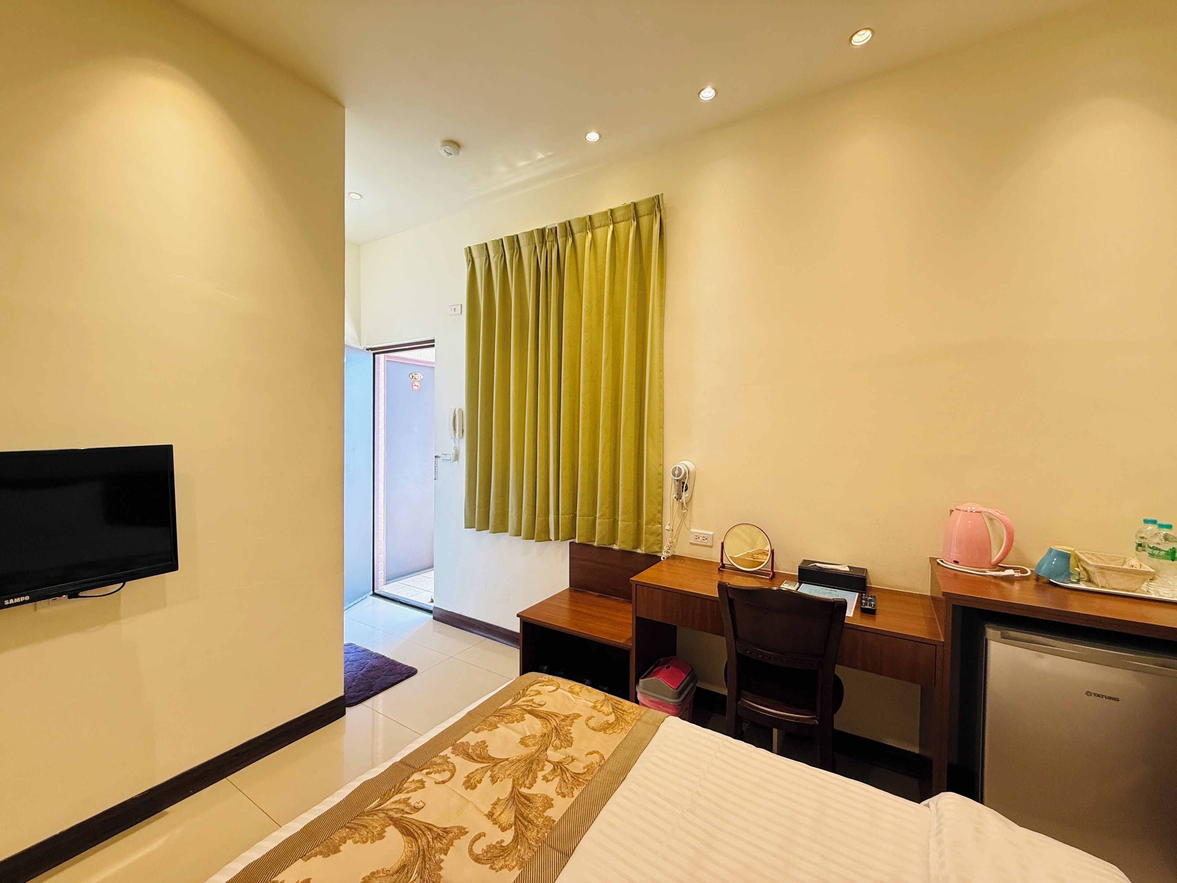 Habitación de hotel con cama, escritorio, silla, televisor, secador de pelo, espejo, hervidor de agua y refrigerador.