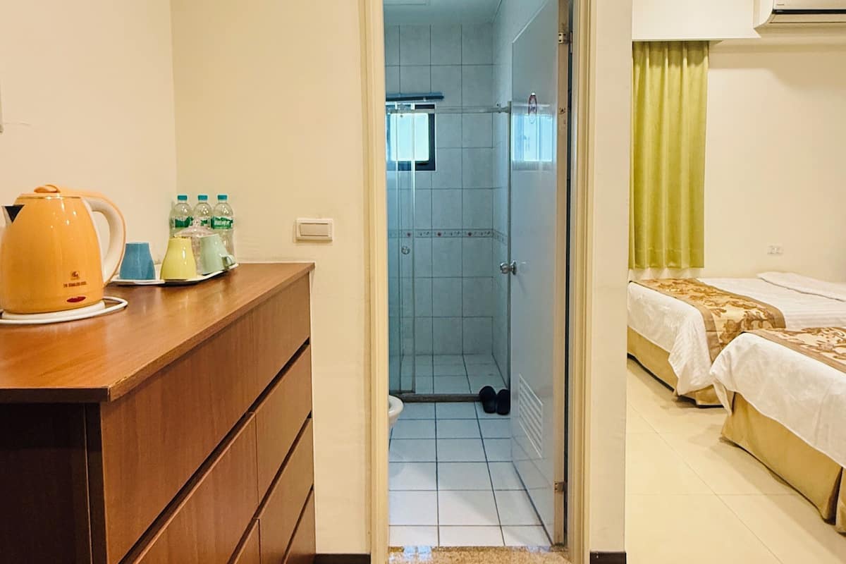 Ein Hotelzimmer mit einem Holzkoffer, einem violetten Teppich, einem Bett mit gemustertem Überwurf und einem sichtbaren Duschbereich durch eine geöffnete Tür.
