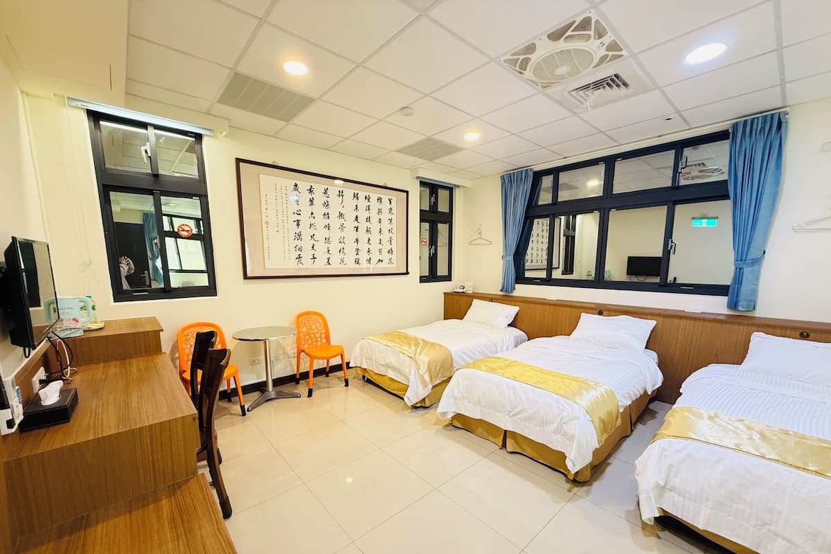 Ein Hotelzimmer mit drei Betten, einem Schreibtisch, einem Sessel und einem großen Fenster.