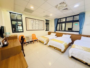 Quarto de hotel com três camas, uma escrivaninha, uma cadeira e uma janela grande.