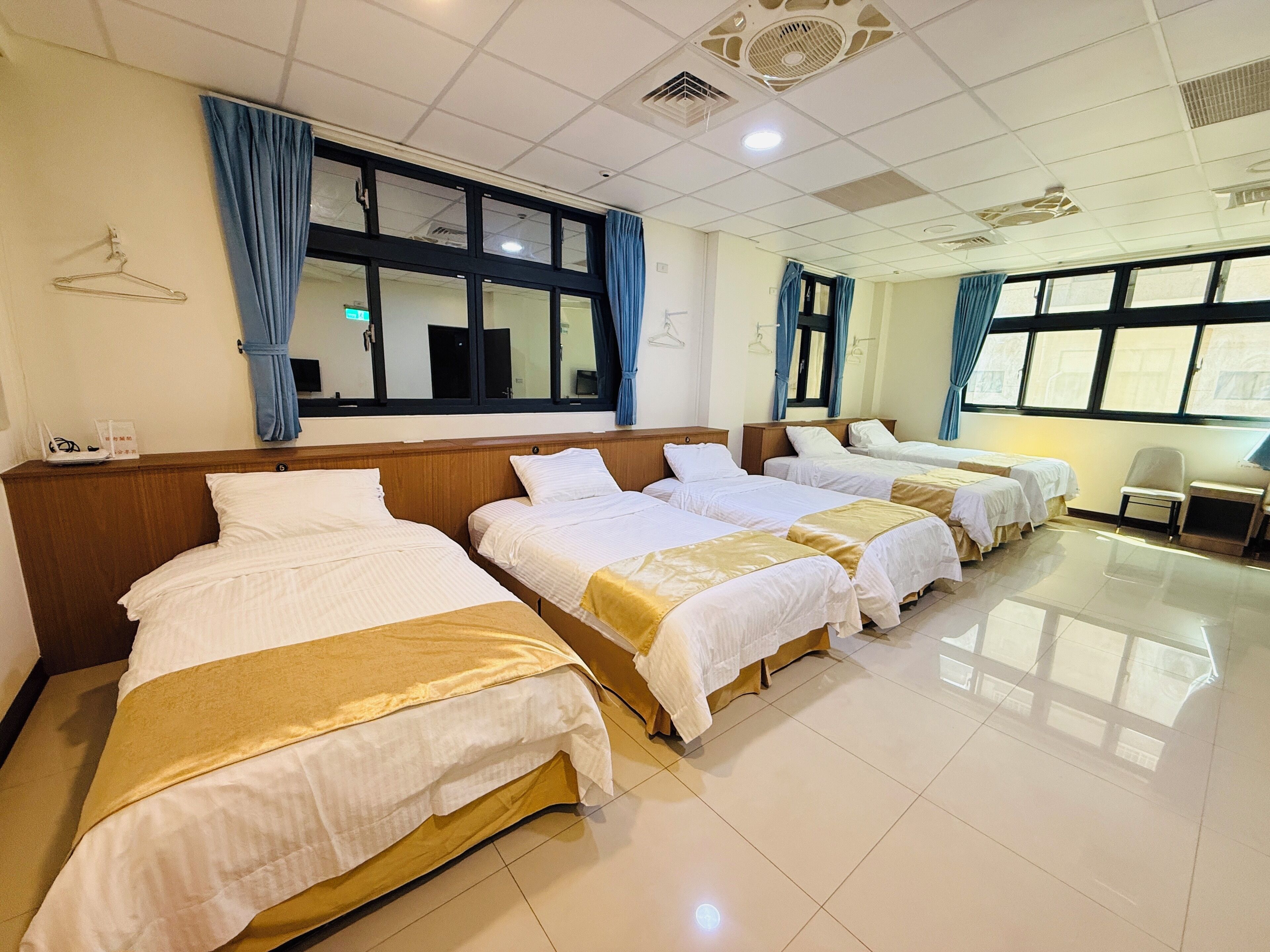 A hotel room with four beds arranged in a row, a desk with a chair, and large windows with curtains.