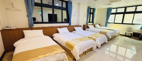 A hotel room with four beds arranged in a row, a desk with a chair, and large windows with curtains.
