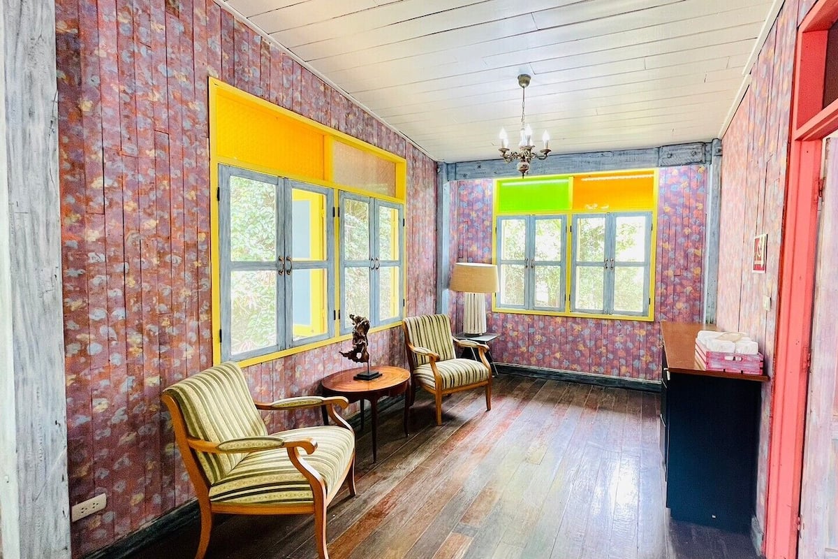 A room with wooden floors, floral wallpaper, and a mix of yellow and green window frames. There are two chairs and a small table with a lamp.