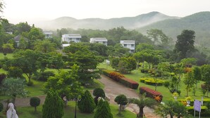 A lush garden with neatly trimmed hedges, a variety of trees, and a clear pathway leading to a row of white buildings. - Baan Ing Khao (Pak Chong)