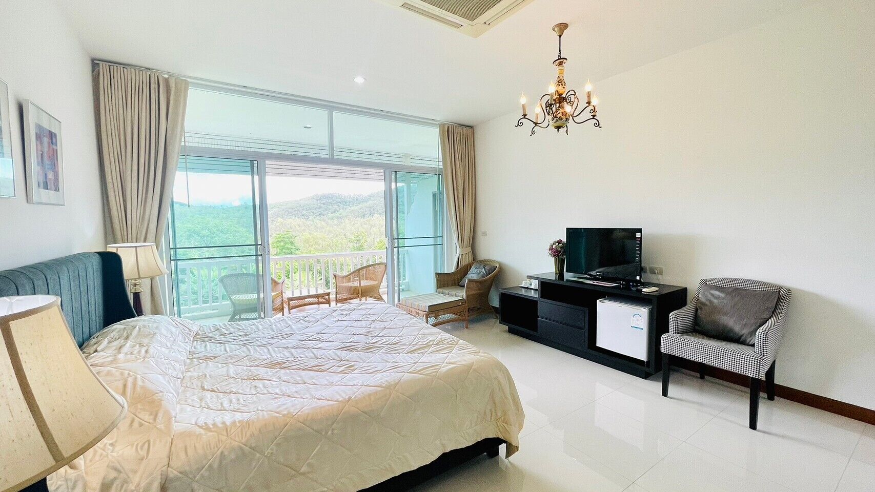 Un dormitorio con una cama grande, televisión, escritorio y silla. Hay un balcón con vista a zonas verdes.