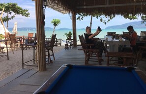 A pool table in a beachside setting with wooden tables and chairs, a tiki bar, and a view of the sea. - Sea Safari (Rawai)