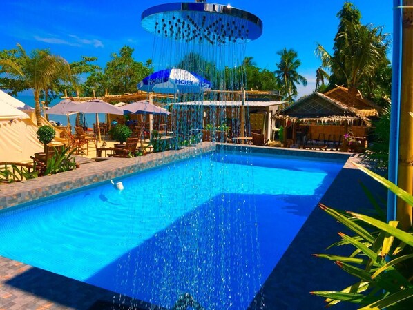 A swimming pool with a water feature, surrounded by lounge chairs and umbrellas, and tropical vegetation. - Sea Safari (Rawai)