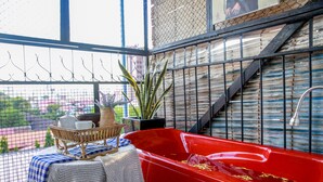 Una bañera roja en una habitación con vistas a través de una ventana con malla metálica, una cesta de mimbre con tazas y una tetera, y una planta en maceta.