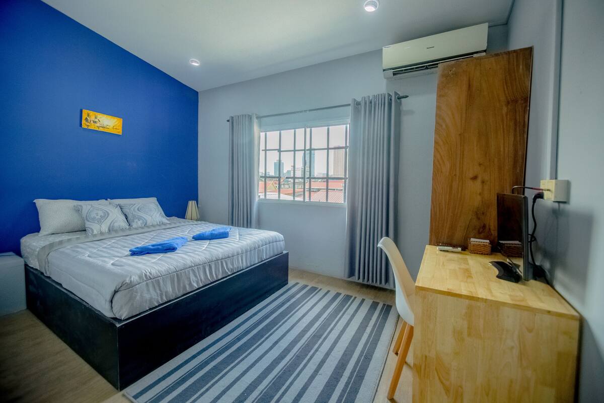Ein Schlafzimmer mit einem Bett, einem Schreibtisch, einem Sessel, einem Fenster mit Vorhängen, einer Klimaanlage und einem Bild an der Wand.