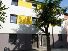 Un edificio moderno con fachada amarilla y blanca, múltiples ventanas y un árbol en primer plano.