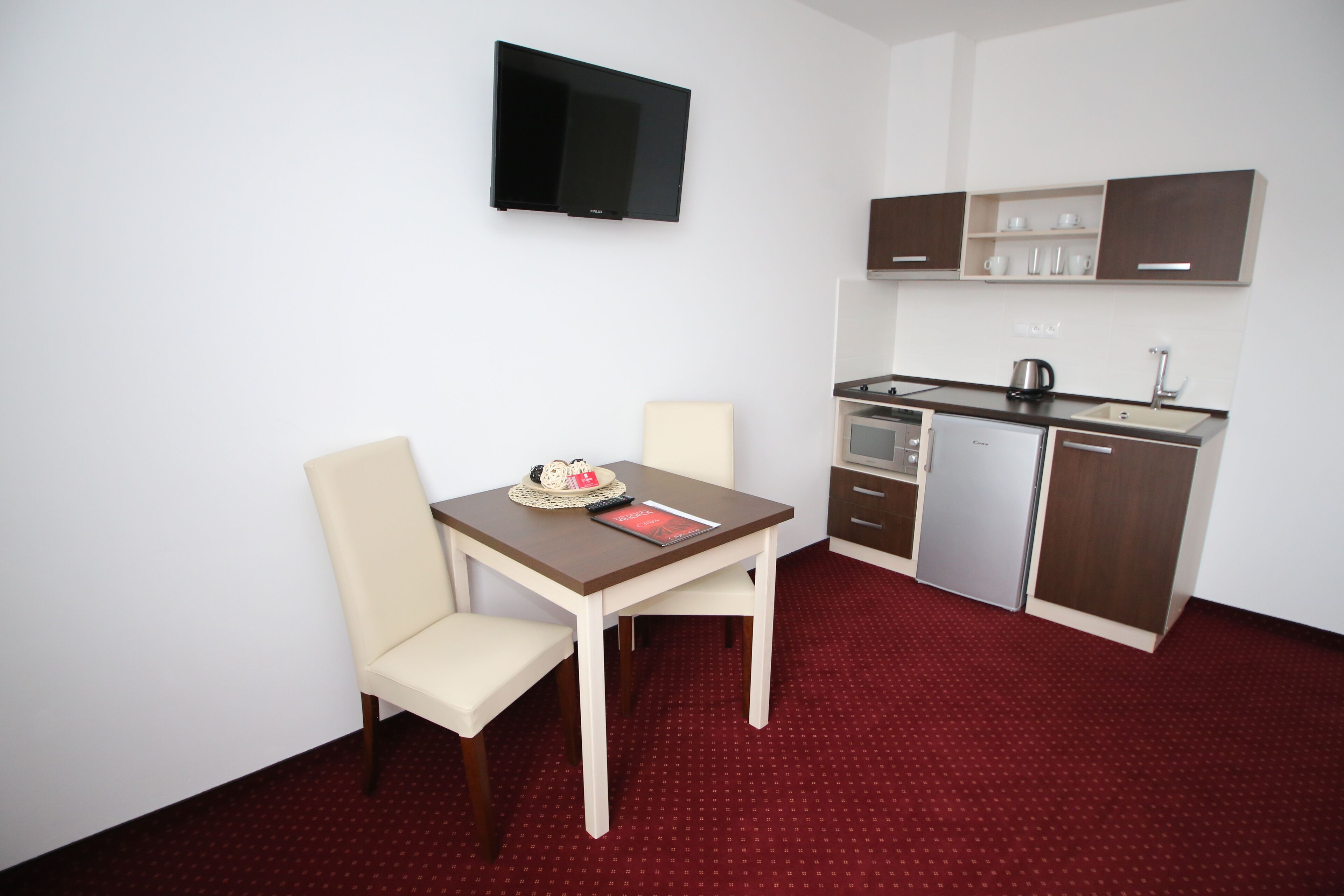 A compact kitchen with a dining table and chairs, a flat-screen TV, and a red carpeted floor.