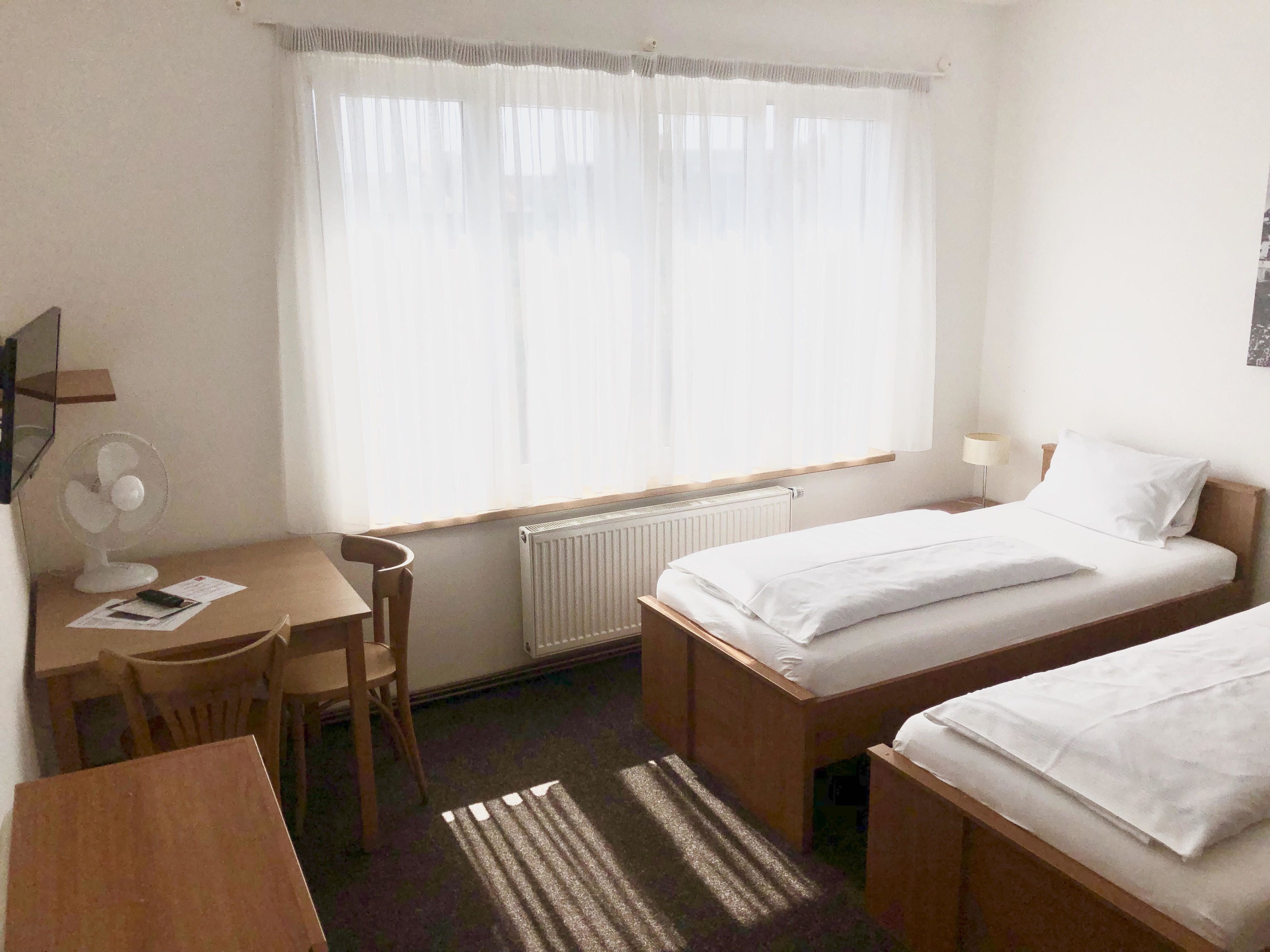 Een hotelkamer met twee eenpersoonsbedden, een bureau met stoel, een radiator en een raam met dunne gordijnen.