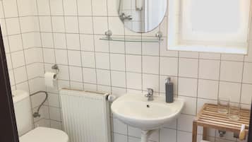 Un baño pequeño con inodoro, lavamanos y espejo. Hay un perchero para toallas y un estante con artículos de aseo.