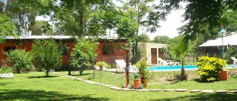 A well-maintained garden with a swimming pool, surrounded by trees and a building with a red roof.