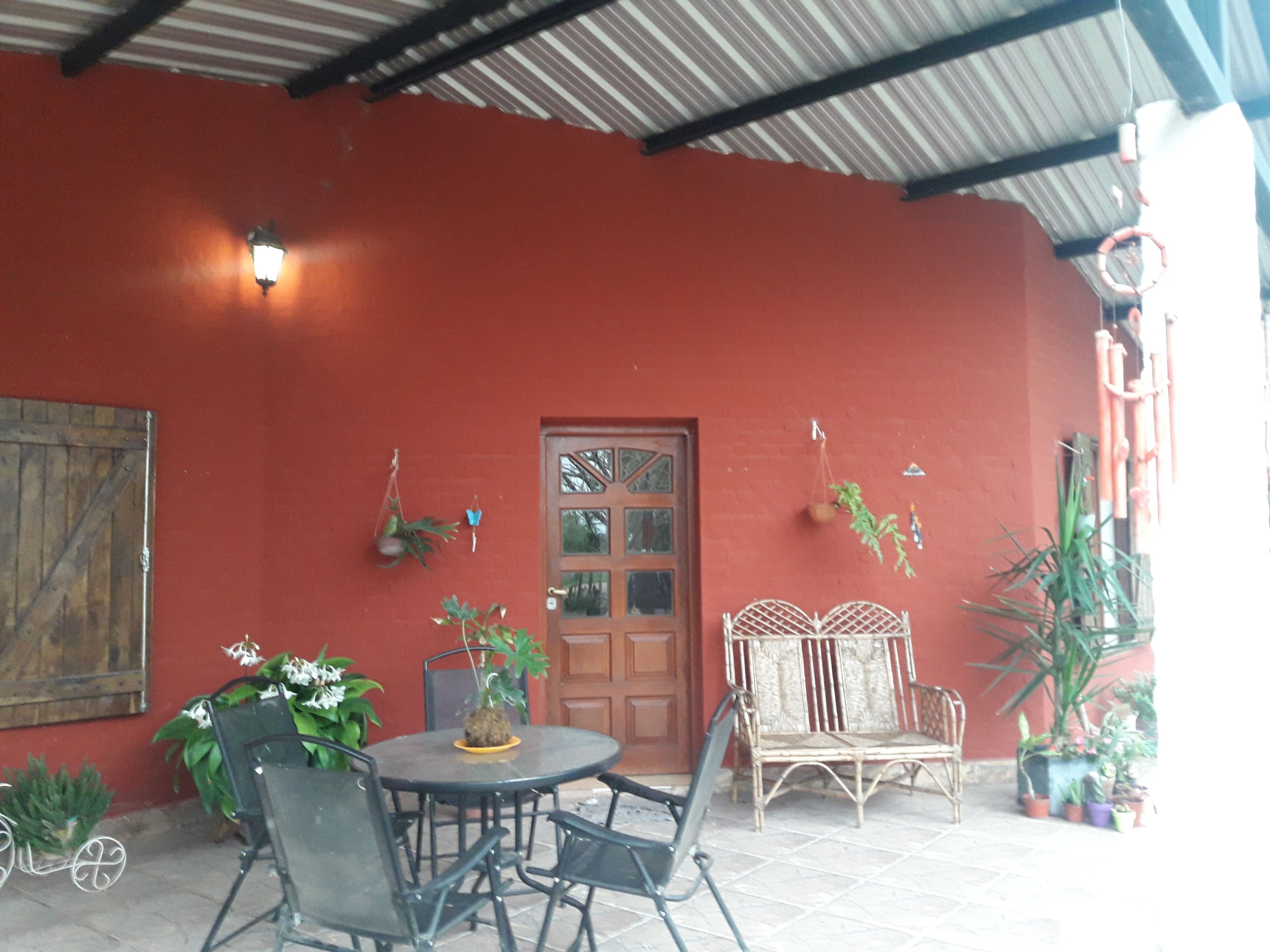 Un patio al aire libre con pared roja, puerta de madera y una mesa redonda con sillas. Hay plantas en macetas y un banco de metal.